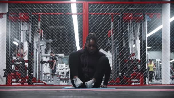 Африкано-американская женщина, сидящая на коврике для йоги в спортзале и греющая свое тело — стоковое видео