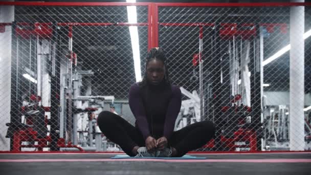 Una mujer afroamericana sentada en una esterilla de yoga en el gimnasio e inclinada hacia adelante — Vídeos de Stock