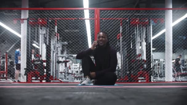 Улыбающаяся афроамериканка сидит на коврике для йоги в спортзале и разговаривает с кем-то по телефону — стоковое видео