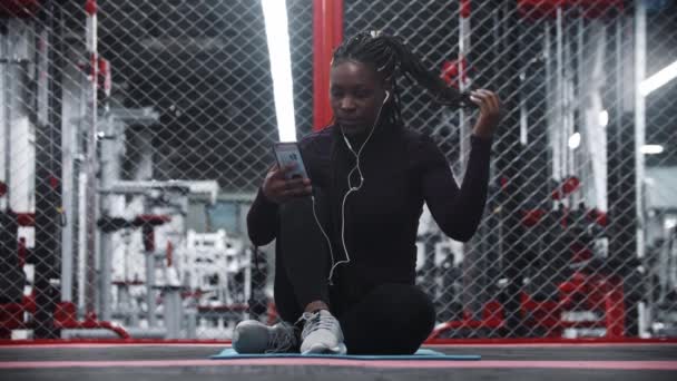 Usmívající se afro-americká žena sedící na podložce jógy v tělocvičně a se selfie na telefonu — Stock video