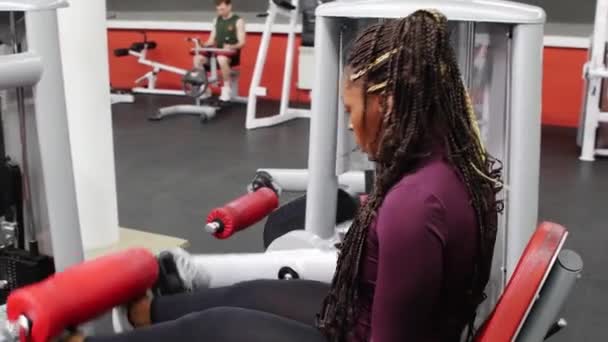 Treinamento de esportes em ambientes fechados - mulher negra com tranças treinando suas pernas em um equipamento de exercício — Vídeo de Stock