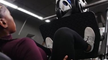 Spor antrenmanı içeride - uzun örgülü siyah bir kadın egzersiz aletleri üzerinde bacaklarını eğitiyor - ağırlık kaldırıyor