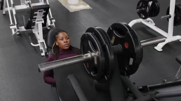 Sporttraining drinnen - schwarze Frau setzt sich auf ein Trainingsgerät und beginnt mit dem Training ihrer Beine - Gewicht mit einer Hantel nach oben schieben — Stockvideo