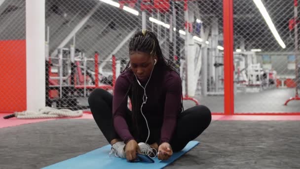 Спорт в тренажерном зале - африканско-американская женщина сидит на коврике для йоги в тренажерном зале и использует свой телефон — стоковое видео
