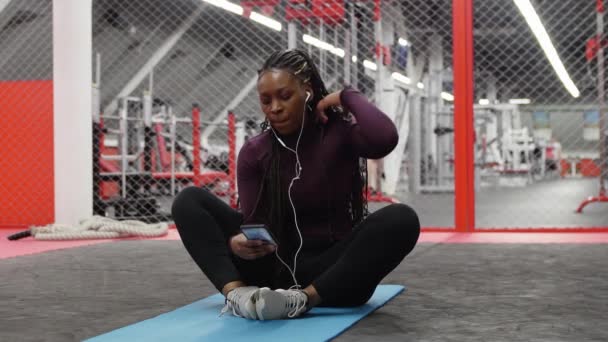 Sport in de sportschool - Afrikaans-Amerikaanse vrouw zit op yoga mat in de sportschool en met behulp van sociale media op haar telefoon — Stockvideo