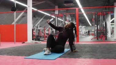 Spor salonunda spor. Afro-Amerikalı bir kadın yoga minderinde oturup selfie çekiyor.