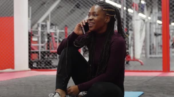 Sport az edzőteremben - afro-amerikai nő ül jóga szőnyegen az edzőteremben, és beszél valakivel a telefonon — Stock videók