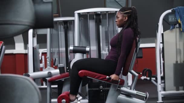 Uma mulher negra trabalhando em um ginásio em um equipamento de exercício de perna — Vídeo de Stock