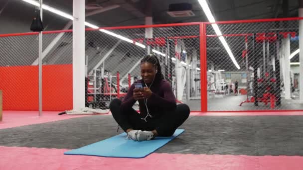 Deportes en el gimnasio - mujer afroamericana sonriente sentada en una esterilla de yoga en el gimnasio y usando las redes sociales en su teléfono — Vídeos de Stock