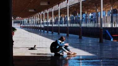 Genç bir gezgin açık hava tren istasyonunda oturuyor ve sokak kedilerinin koşuşunu izliyor.