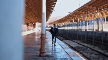 Güneş gözlüklü genç bir adam açık hava tren istasyonunda yürüyor.