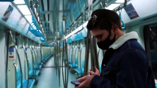 Mladý muž v černé masce stojí v prázdném vlaku metra a dívá se na svůj telefon — Stock video