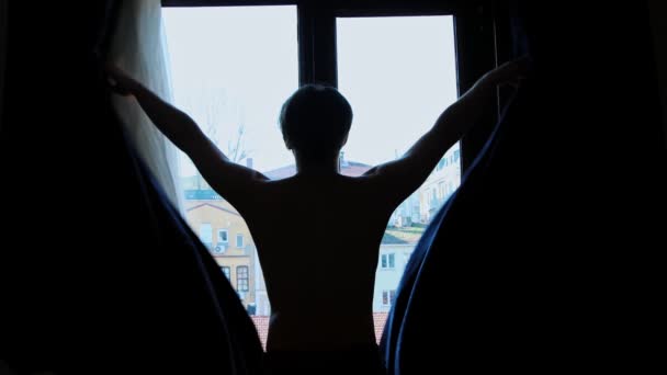 Молодий голий чоловік відкриває віконні штори вранці і дивиться у вікно — стокове відео