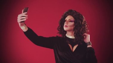 Kırmızı arka planda abartılı elbiseler giyen genç bir adam selfie çekiyor ve gülümsüyor.
