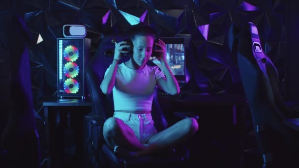 女の子のゲーマーは、ゲームクラブで椅子に座って、彼女のヘッドフォンを置く-ロリポップを吸う — ストック動画