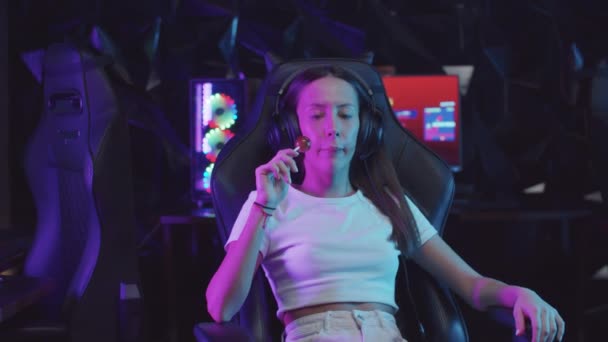 Девушка-геймер сидит в кресле в игорном клубе и сосет леденец — стоковое видео