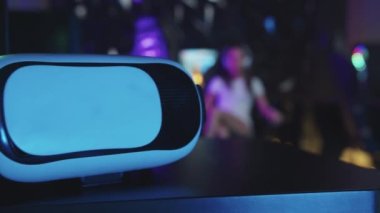 Neon oyun kulübünde sandalyede oturan genç oyuncu kadın. Ön planda VR gözlükleri.