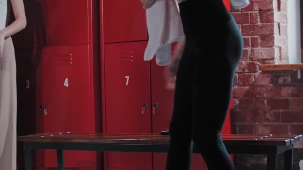 Two young women sits down on a bench in the locker room after training and about to change their clothes — Stock Video