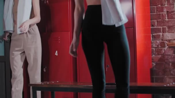 Two young women sits down on a bench in the locker room after training and takes off the shoes — Stock Video