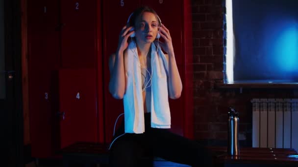 Deportes - mujer bonita joven sentada en el vestuario con una toalla y auriculares en el cuello — Vídeos de Stock