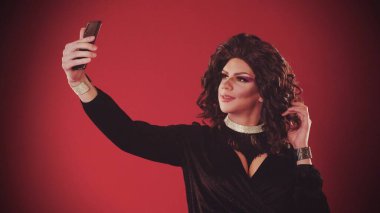 Kırmızı arka planda abartılı elbiseler giyen genç bir adam selfie çekiyor ve gülümsüyor.