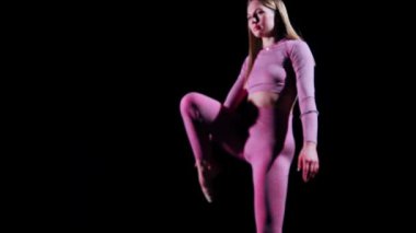 Jimnastik. Pembe kostümlü genç bir kadın karanlık stüdyoda yürüyor ve bacağını kaldırıyor.