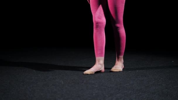 Γυμναστική - νεαρή γυναίκα με ροζ στολή περπατάει στο σκοτεινό στούντιο και σηκώνει το πόδι της ψηλά — Αρχείο Βίντεο