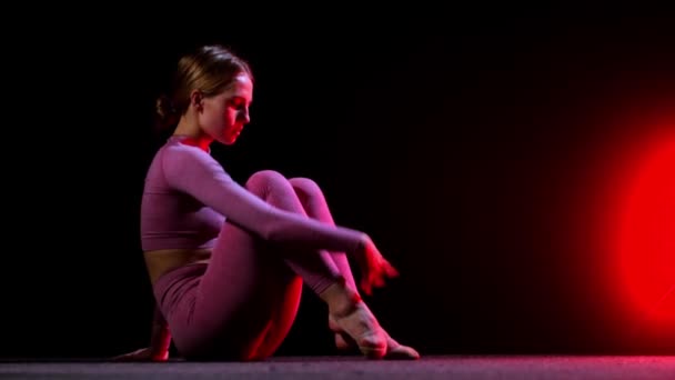 Gimnasia - joven mujer de plástico en traje rosa sentada en el suelo y levantando la pierna mostrando su estiramiento - iluminación de neón rojo — Vídeo de stock
