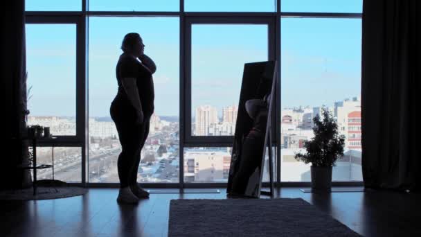 A big overweight woman stands in the office with panoramic windows and looks at herself in the mirror — Stock Video