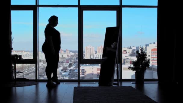 An overweight woman stands in the office with panoramic windows and looks at herself in the mirror — Stock Video