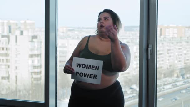 Vermoeide vrouw met overgewicht in sportkleding gooit een naambordje weg met een bordje VROUW KRACHT en begint vrolijk te dansen — Stockvideo