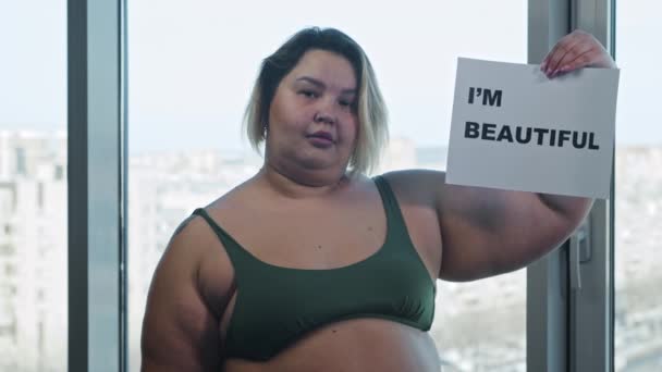 Een vrouw met overgewicht die een naamplaatje draagt met een bord IM BEAUTIFUL en een hamburger eet — Stockvideo