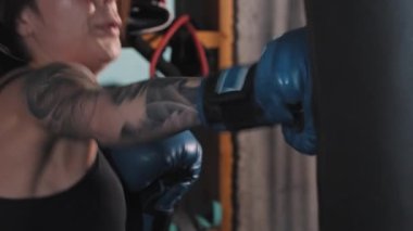 Spor salonunda boks yaparken dövmeli bir kadın boksör eldiveni giyerek kum torbasını yumrukluyor.