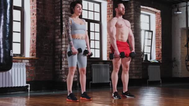 Un par de mujeres tatuadas y un hombre en forma en el gimnasio sosteniendo pesas y inclinándose hacia abajo para el entrenamiento de manos — Vídeo de stock