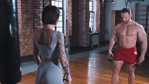 Athletic man and tattooed woman training in the gym together - doing hands exercises with dumbbells one at a time — Stock Video