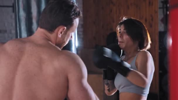 Боксерская тренировка в тренажерном зале - женщина боксер тренирует свои удары по боксерской груше с ее тренером мужского пола, держащим ее — стоковое видео