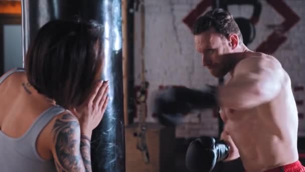 Boxning i gymmet - man boxare tränar sina slag på boxningssäck med sin kvinnliga tränare håller den — Stockvideo