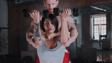 Çekici, gülümseyen bir kadın ellerini dambıllarla şişiriyor. Eğitmeni onu izliyor ve ona yardım ediyor.