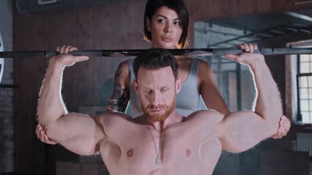 Atraktivní pár trénink v tělocvičně - atletický muž pumpovat svaly rukou s činkou a tetovaná žena stojí za ním a drží ho pod lokty — Stock video