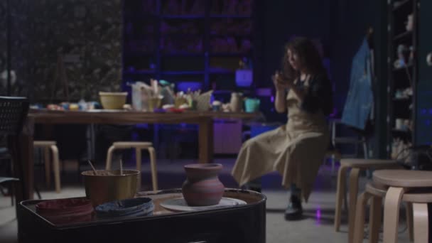 Um pote novo fresco girando na roda e uma jovem sentada à mesa com barro nas mãos — Vídeo de Stock