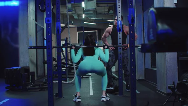 Gym training - woman trains her butt with dumbbell on her shoulders under the supervision of a trainer — Stok fotoğraf