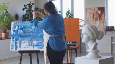 Bir kadın ressam telefonundaki yağlı boya tablosunun fotoğraflarını çekiyor.