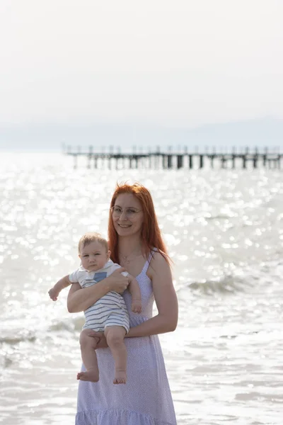 Madre feliz de vacaciones - mujer sonriente de pie en la playa sosteniendo a su pequeño hijo —  Fotos de Stock
