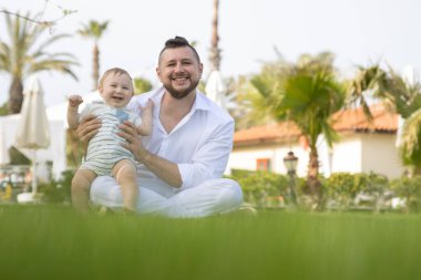 Mutlu baba ve oğlu çimlerin üzerinde oturup kameraya bakıyorlar.