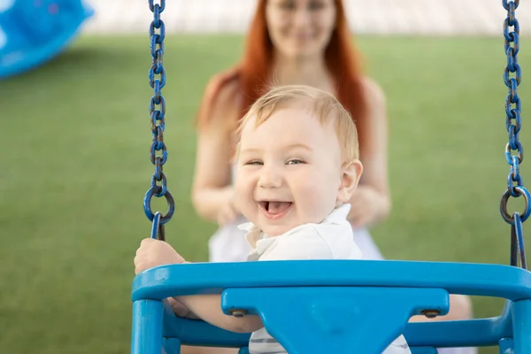 Egy kis nevető baba hintázik egy külső játszótéren a vörös anyjával. — Stock Fotó