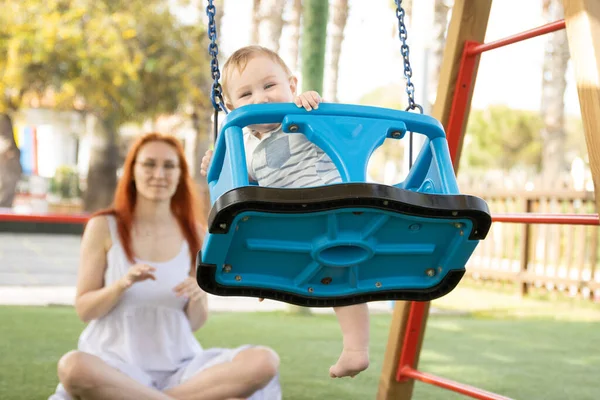 Egy kis vicces baba hintázik egy külső játszótéren a vörös anyjával. — Stock Fotó