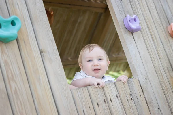Bebé divertido se sienta en una casa de madera en el patio de recreo — Foto de Stock