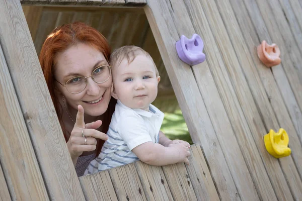 Маленька дитина і його мати сидять в дерев'яному будинку на дитячому майданчику дивлячись в камеру — стокове фото
