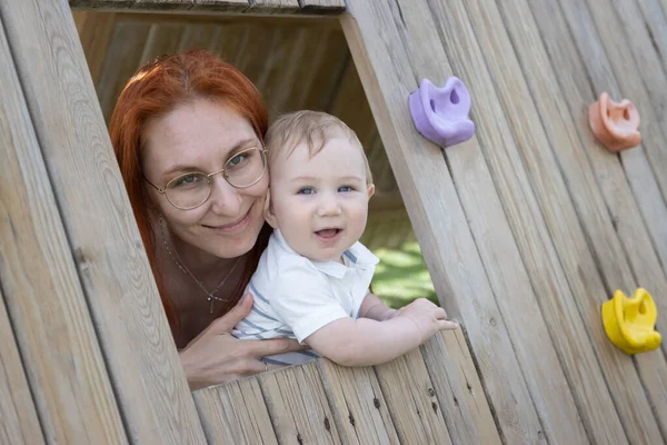 Lachende baby en zijn moeder zit in een houten huis op de speelplaats - kijken in de camera — Stockfoto