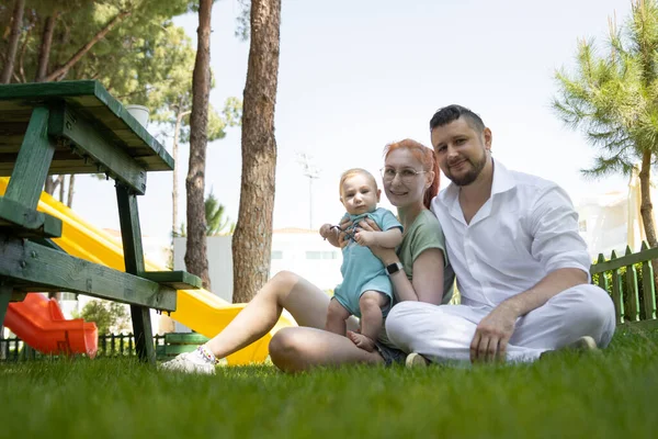 Los padres felices y su lindo hijo se sientan en la hierba mirando a la cámara —  Fotos de Stock